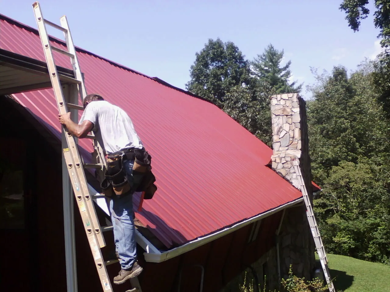 Roofer on a ladder beside a red metal roof and stone chimney — Soffit & Fascia Repair by Roofing Brothers in Newton Hamilton
